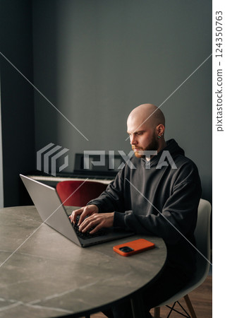 Vertical portrait of young bald programmer with beard and ear piercing sitting at round table in home office, concentrating on work on laptop computer looking to screen, mobile phone nearby. 124350763
