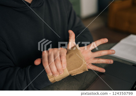 Close-up cropped shot of male hands wearing wrist brace, massaging painful wrist while working remotely from home office, surrounded by laptop and notebook. Concept of struggles of remote work 124350799