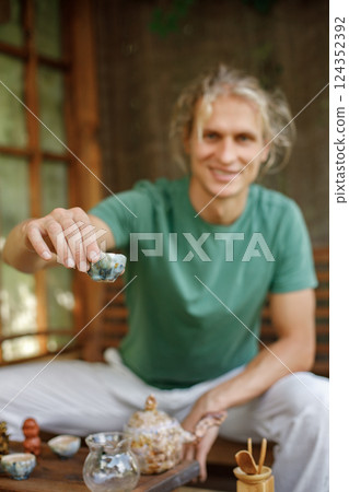 Blonde man preparing for a tea ceremony. Man sitting in outdoor Japanese spring garden in backyard porch of zen home. Focus on a cup in man`s hands. Blonde man preparing for a tea ceremony. Man sitting in outdoor Japanese spring garden in backyard porch of zen home. Focus on a cup in man`s hands. 124352392