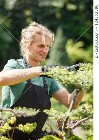 Blonde caucasian man planting flowers in pot with garden tools. Man wearing green t-shirt and black apron. Man spending peaceful morning in garden hear house. Blonde caucasian man planting flowers in pot with garden tools. Man wearing green t-shirt and black apron. Man spending peaceful morning in garden hear house. 124352396