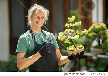 Blonde caucasian man planting flowers in pot with garden tools. Man wearing green t-shirt and black apron. Man spending peaceful morning in garden hear house. 124352397