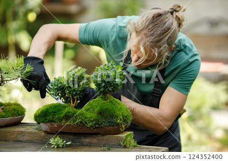 Blonde caucasian man planting flowers in pot with garden tools. Man wearing green t-shirt and black apron. Man spending peaceful morning in garden hear house. 124352400