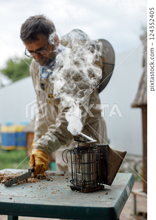 Experienced senior apiarist in his apiary setting a fire in a bee smoker. Beekeeper harvesting honey. Bee smoker is used to calm bees before frame removal. 124352405