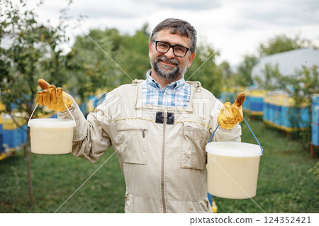 Beekeeper holding bucket of honey in hands. Farmer wearing bee suit working with honeycomb in apiary. Man looking at camera. 124352421