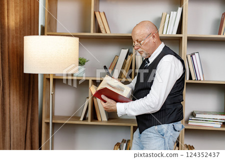 Senior man standing near bookshelves and reading book at home. Man using a magnifying glass. Man wearing white shirt and black waistcoat. 124352437