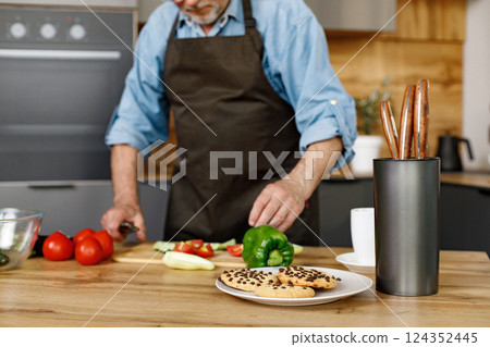 Retired senior man cooking from green salat, pepper and tomatoes. Man standing in kitchen interior. Man wearing brown apron and shirt. Retired senior man cooking from green salat, pepper and tomatoes. Man standing in kitchen interior. Man wearing brown apron and shirt. 124352445