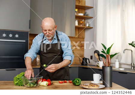 Retired senior man cooking from green salat, pepper and tomatoes. Man standing in kitchen interior. Man wearing brown apron and shirt. Retired senior man cooking from green salat, pepper and tomatoes. Man standing in kitchen interior. Man wearing brown apron and shirt. 124352446