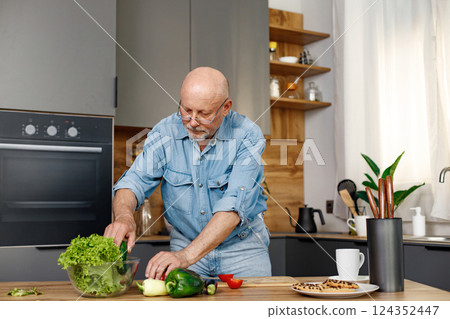 Retired senior man cooking from green salat, pepper and tomatoes. Man standing in kitchen interior. Man wearing jeans and shirt. 124352447