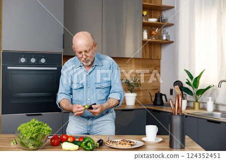Retired senior man cooking from green salat, pepper and tomatoes. Man standing in kitchen interior. Man wearing jeans and shirt. Retired senior man cooking from green salat, pepper and tomatoes. Man standing in kitchen interior. Man wearing jeans and shirt. 124352451