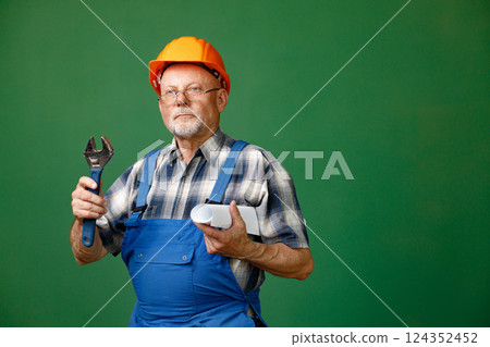 Studio portrait of a man isolated on green background. Seniour man worker in a blue plaid shirt and blue overall holds a straight pipe wrench or spanner. Man wearing eyeglasses and orange helmet. Studio portrait of a man isolated on green background. Seniour man worker in a blue plaid shirt and blue overall holds a straight pipe wrench or spanner. Man wearing eyeglasses and orange helmet. 124352452