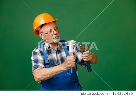 Studio portrait of a man isolated on green background. Seniour man worker in a blue plaid shirt and blue overall holds a straight pipe wrench or spanner. Man wearing eyeglasses and orange helmet. 124352459