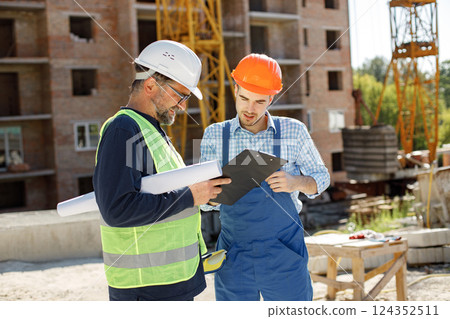 Worker and architect watching some details on a construction. Two engineers talking at building site with construction structure in background. Men wearing helmets. 124352511