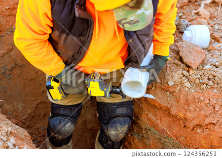 Construction worker in orange hoodie, gloves uses power tool to cuts pvc wastewater pipes in trench 124356251