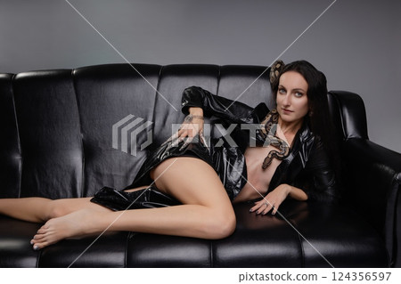 Woman posing elegantly with a snake on a sleek black couch in seductive lingerie Woman posing elegantly with a snake on a sleek black couch in seductive lingerie 124356597