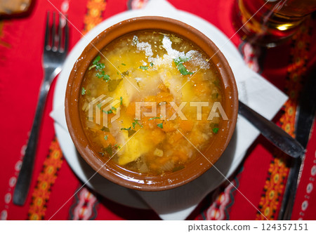 Deep clay bowl with Bulgarian fish and vegetable soup, complemented by cutlery and strong drink 124357151