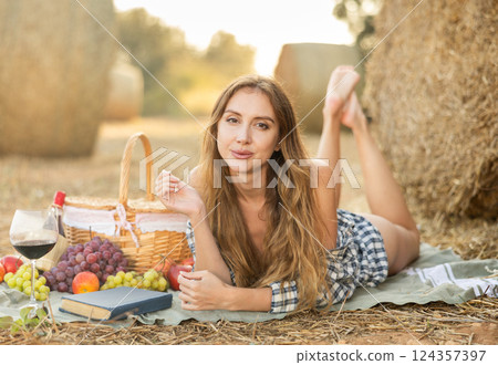 Beautiful cowboy woman in checked shirt lying down and posing near haystacks with basket of fruit and wine, fashion concept Beautiful cowboy woman in checked shirt lying down and posing near haystacks with basket of fruit and wine, fashion concept 124357397