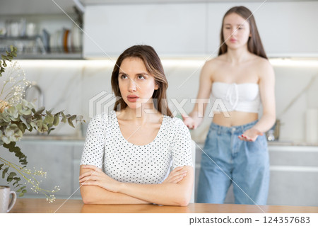 Angry mother sitting at the kitchen table, daughter standing behind Angry mother sitting at the kitchen table, daughter standing behind 124357683