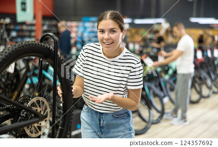 Positive young girl looking for and trying wheel new bike in sports bicycle store Positive young girl looking for and trying wheel new bike in sports bicycle store 124357692