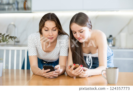 Two young women talking while looking at phones in kitchen Two young women talking while looking at phones in kitchen 124357773