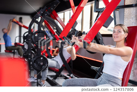 Girl doing strength training in gym, exercising upper body muscles on shoulder press machine 124357806