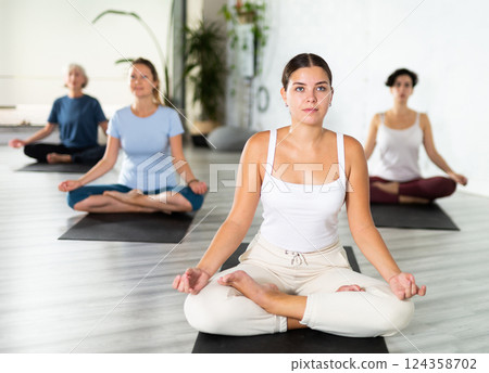 Relaxed young European female sitting in meditation Lotus Pose Padmasana during yoga class with other women Relaxed young European female sitting in meditation Lotus Pose Padmasana during yoga class with other women 124358702