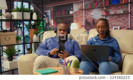 Young woman working on freelance tasks while her partner browses phone, modern couple using their devices sitting on the couch in the living room. People enjoying time at home. Camera A. 124360225