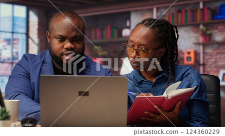 Black remote employees organizing calendar appointments using laptop and ledger, checking papers and synchronizing important tasks on schedule. Man and woman review files at home. Camera B. 124360229
