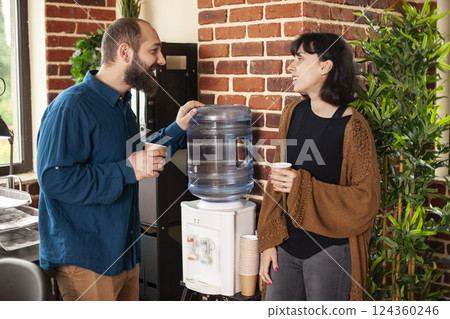Bearded man and female colleague hold paper cups standing near brick wall and exchanging business project ideas. Businesswoman and male coworker stand next to water dispenser taking break at office. 124360246