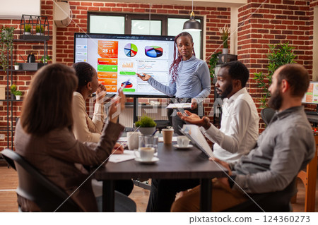 Black female analyst points to screen displaying business analytics, giving explanations to diverse employees seated at table. Businesswoman with clipboard, leading team meeting in brick wall office. Black female analyst points to screen displaying business analytics, giving explanations to diverse employees seated at table. Businesswoman with clipboard, leading team meeting in brick wall office. 124360273