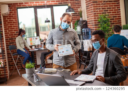 Caucasian man shares data on a tablet during a conversation with his black colleague in a safe office setting. Male coworkers collaborate at a desk, both masked in brick wall workplace. Caucasian man shares data on a tablet during a conversation with his black colleague in a safe office setting. Male coworkers collaborate at a desk, both masked in brick wall workplace. 124360295