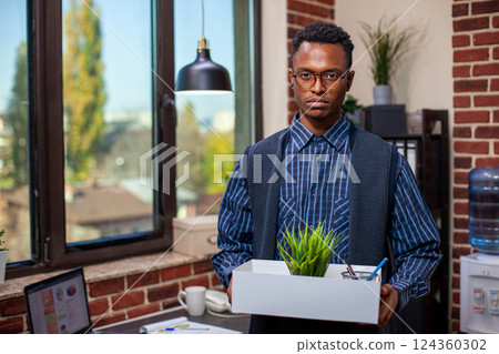 Portrait of black male employee holds a box containing green plant and stationery, standing in brick wall workspace. Businessman with a white carton of office materials, looking at camera. Portrait of black male employee holds a box containing green plant and stationery, standing in brick wall workspace. Businessman with a white carton of office materials, looking at camera. 124360302