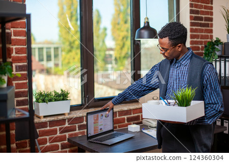 Businessman carrying work belongings and closing his laptop on desk, preparing to leave the office. Professional manager with box in hand, ready to end the day, shutting off his personal computer. Businessman carrying work belongings and closing his laptop on desk, preparing to leave the office. Professional manager with box in hand, ready to end the day, shutting off his personal computer. 124360304