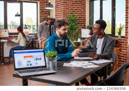 Male entrepreneurs brainstorming business ideas and reviewing company data on a tablet in modern workplace. Young men seated at desk with shared device, analyzing annual reports collaboratively. 124360306