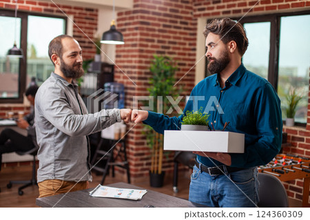 Departing male employee holds a box of belongings and exchanges a fist bump with a colleague, in brick wall workplace. Professional farewell moment between caucasian businessmen in modern office. 124360309