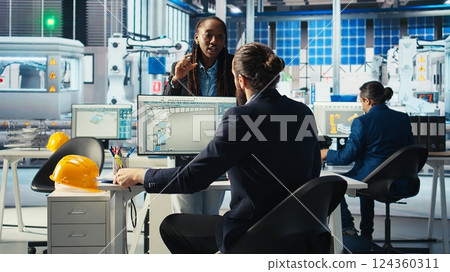 Photovoltaics factory production manager looking over schematics, instructing worker on adjustments to make. Solar panels plant engineer looking over assembly lines blueprints, talking with employee 124360311