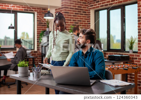 Black businesswoman with a clipboard discusses marketing strategy ideas with male colleague in brick wall workspace. Caucasian bearded man listens intently to female coworker at his desk in office. Black businesswoman with a clipboard discusses marketing strategy ideas with male colleague in brick wall workspace. Caucasian bearded man listens intently to female coworker at his desk in office. 124360343