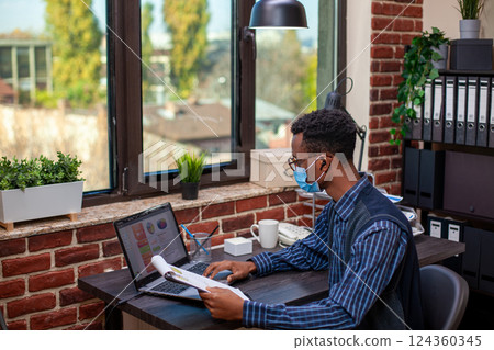 Male analyst with clipboard, researching financial indicators on laptop and planning business presentation. Black man seated in brick wall office is comparing company strategy ideas with documents. Male analyst with clipboard, researching financial indicators on laptop and planning business presentation. Black man seated in brick wall office is comparing company strategy ideas with documents. 124360345