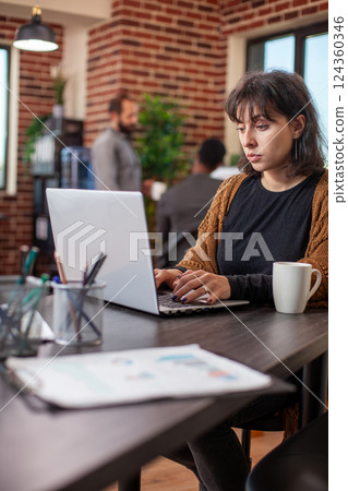 Portrait of female manager focused on her laptop, preparing business project plan in brick wall office. Professional woman sits at desk with coffee, browsing for marketing strategies on digital device Portrait of female manager focused on her laptop, preparing business project plan in brick wall office. Professional woman sits at desk with coffee, browsing for marketing strategies on digital device 124360346