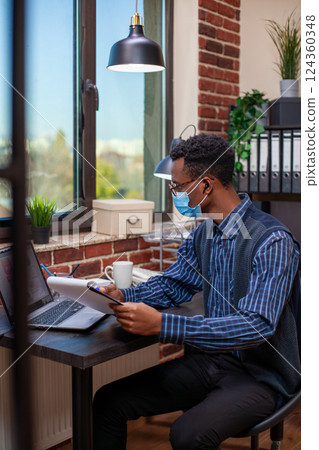 Male analyst with face mask, listening to music with earphones and reading company research information at desk near window. Entrepreneur holds clipboard, analyzing business project plans on laptop. Male analyst with face mask, listening to music with earphones and reading company research information at desk near window. Entrepreneur holds clipboard, analyzing business project plans on laptop. 124360348