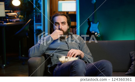 Man at home looking in disbelief at thriller film on TV, stuffing mouth with popcorn. Stupefied person at home watching suspenseful movie on VOD channel, perplexed by plot twist, camera A Man at home looking in disbelief at thriller film on TV, stuffing mouth with popcorn. Stupefied person at home watching suspenseful movie on VOD channel, perplexed by plot twist, camera A 124360361
