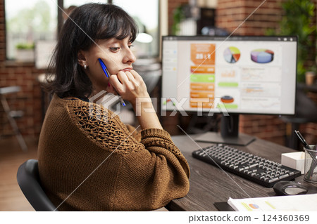 Businesswoman sits overwhelmed, staring at desk filled with financial paperwork in startup office. Female analyst holding her chin and looking tired after reviewing marketing strategy on computer. 124360369