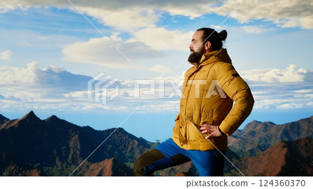 Hiker pauses to admire the panoramic mountain scenery on the hill top, endless sky and rugged peaks creating a breathtaking backdrop for mental clarity. Adventure seeker. Camera B. 124360370