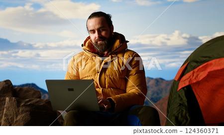 Workaholic explorer always connected and addicted to uninterrupted work, opening his laptop on the mountain top. Young hiker with internet dependency enjoys strong signal. Camera B. 124360371