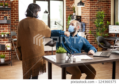 Young man and woman in business casual clothing greet each other with elbow bump at desk in brick wall room. Caucasian colleagues wearing face masks and ensuring pandemic safety in startup office. 124360375