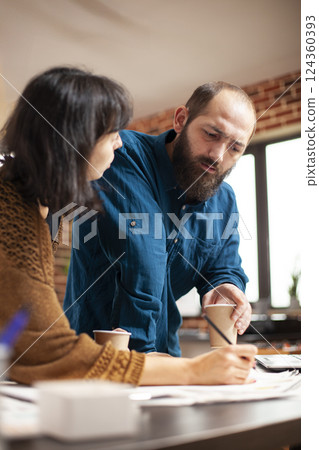 Bearded man holding coffee cup and providing input during business meeting with female analyst at office desk. Closeup on caucasian coworkers having warm beverage and reviewing financial paperwork. Bearded man holding coffee cup and providing input during business meeting with female analyst at office desk. Closeup on caucasian coworkers having warm beverage and reviewing financial paperwork. 124360393