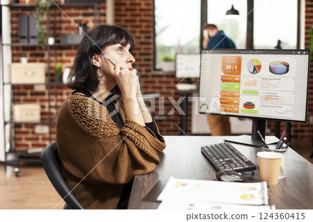 Female employee sits at office desk having brainstorming session as desktop monitor displays market data. Woman with pen, deeply thinking about project ideas for startup company at brick wall room. Female employee sits at office desk having brainstorming session as desktop monitor displays market data. Woman with pen, deeply thinking about project ideas for startup company at brick wall room. 124360415