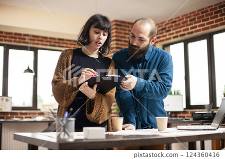 Caucasian male and female colleagues looking at company research data on clipboard. Bearded man and young woman standing at office desk and reviewing financial paperwork in brick wall room. 124360416