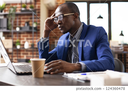 Male employee brainstorming ideas and using his personal computer at office desk. African american businessman sits thoughtfully with hand on forehead, analyzing marketing strategy trends on laptop. 124360478