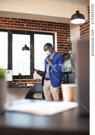Black male entrepreneur with face mask, reviewing company strategies on his digital device. Businessman holding his glasses and analyzing marketing opportunities on laptop in brick wall office. Black male entrepreneur with face mask, reviewing company strategies on his digital device. Businessman holding his glasses and analyzing marketing opportunities on laptop in brick wall office. 124360509