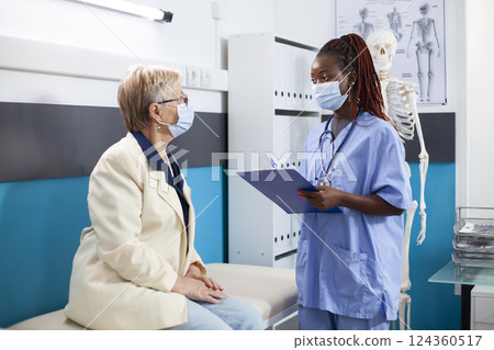 African American nurse with clipboard stands by hospital bed discussing with senior white woman as both wear face masks. Retired patient listens attentively to young medic during medical appointment. African American nurse with clipboard stands by hospital bed discussing with senior white woman as both wear face masks. Retired patient listens attentively to young medic during medical appointment. 124360517